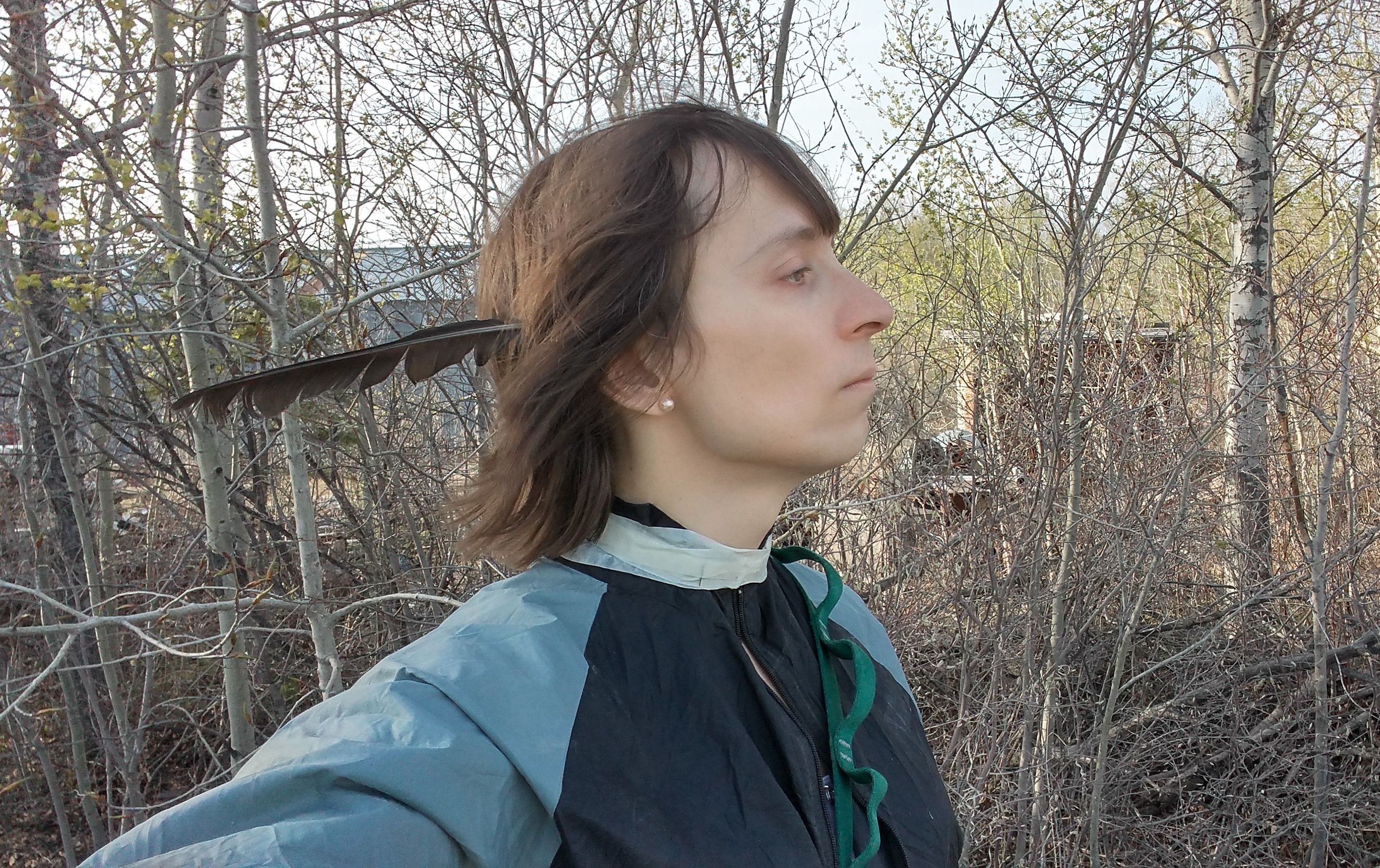 KatieLynne, in a forest, with a feather in her ear, and tape around her neck, it's to keep the woodticks out. But yes, standing in a forest, a kinda selfie from the side type of thing.